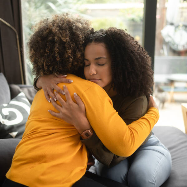 An African-American female psychotherapist comforts the teenage female patient by hugging her. Both of them have the afro hairstyle.